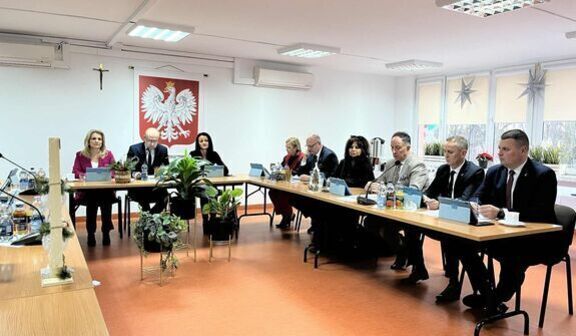 Na zdjęciu widać grupę osób siedzących przy dwóch stołach ustawionych w kształcie litery L. W tle znajduje się godło Polski na ścianie. Pomieszczenie jest jasne i ozdobione roślinami.