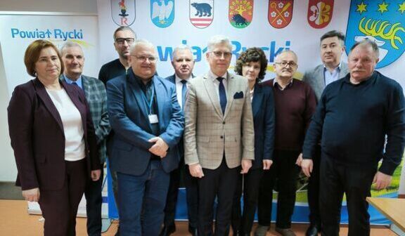 Na zdjęciu znajduje się grupa osób stojących razem na tle tablicy z herbami gmin powiatu ryckiego. Widać mężczyzn i kobietę w formalnych strojach. Atmosfera jest oficjalna.