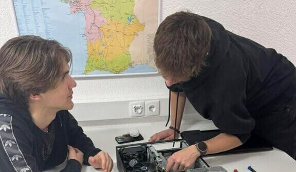 Dwóch młodych mężczyzn pracuje nad otwartym komputerem na stole pełnym narzędzi. Na ścianie za nimi wisi mapa. Oboje są skupieni na zadaniu. Na stole leżą różne śrubokręty i inne narzędzia.