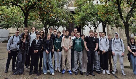 Grupa ludzi pozuje przed ozdobnym, gotyckim budynkiem otoczonym drzewami owocowymi. Budowla ma bogato zdobioną fasadę z detalami rzeźbiarskimi i dużym oknem rozetowym.