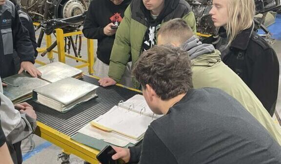 Grupa młodych ludzi stoi wokół stołu w hangarze lub warsztacie lotniczym, przeglądając dokumenty. W tle widać części samolotu oraz śmigło. Uczestnicy są skupieni i zaangażowani w analizę materiałów.