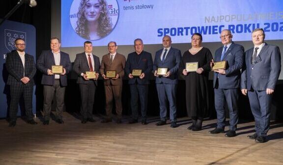 Dziewięć osób stoi na scenie trzymając nagrody w postaci tabliczek. Za nimi na ekranie widnieje napis "Najpopularniejszy Sportowiec Roku 2021". W tle widoczny jest niebieski plakat i zdjęcie kobiety.