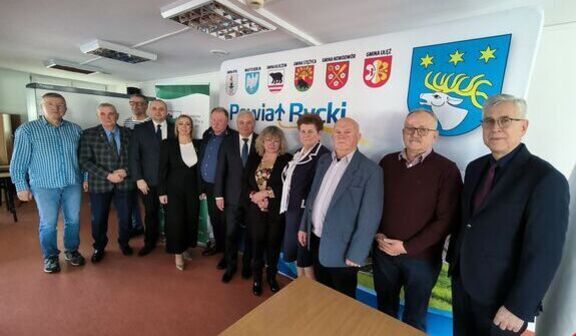 Grupa osób pozuje do zdjęcia w biurze. W tle widoczne są flagi i logo powiatu puckiego. Wszyscy są elegancko ubrani, stoisko w centralnej części zdjęcia.