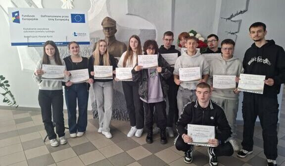 Grupa młodych ludzi stoi w pomieszczeniu z certyfikatami w rękach. Za nimi widoczna jest wykonana z ciemnego materiału rzeźba oraz tablica z logotypami. W tle szare ściany z dekoracjami.