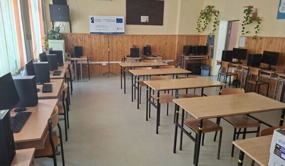 Klasa komputerowa z kilkoma rzędami drewnianych ławek i krzeseł. Na biurkach rozmieszczone są monitory komputerowe. Ściany ozdobione są roślinami i tablicami informacyjnymi.