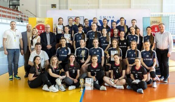 Na zdjęciu grupa siatkarek w ciemnych strojach pozuje z trenerami i oficjelami. Kilka osób trzyma puchary i dyplomy w hali sportowej. W tle widoczne są bannery i trybuny.