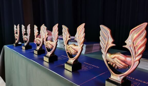 Rząd statuetek z motywem dłoni uścisku stoi na stole, każda na granatowej podstawie. W tle stosy dokumentów. Scena sugeruje przygotowanie do ceremonii wręczenia nagród.