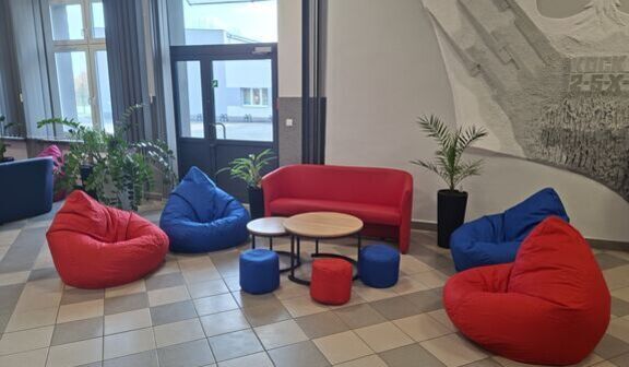 Przytulna przestrzeń z kolorowymi pufami w kształcie worków: czerwonymi i niebieskimi. Na środku okrągły stolik z trzema małymi pufami. W tle ściana z dekoracyjną grafiką i rośliny doniczkowe.