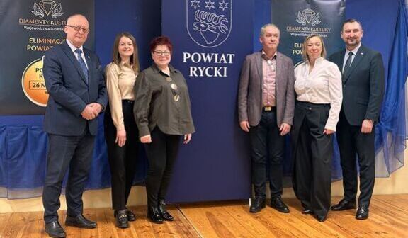 Na zdjęciu grupka ludzi stoi w sali. W tle znajduje się duży baner z napisem "Powiat Rycki". Dookoła są dwa mniejsze banery z hasłem dotyczącym kultury. Podłoga jest drewniana.