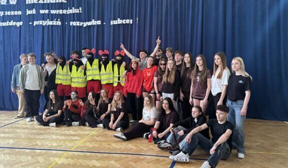 Grupa osób pozuje na sali gimnastycznej. Niektórzy mają na sobie kolorowe stroje, odblaskowe kamizelki i zabawne akcesoria. Za nimi widoczna jest niebieska kurtyna z napisem w języku polskim.