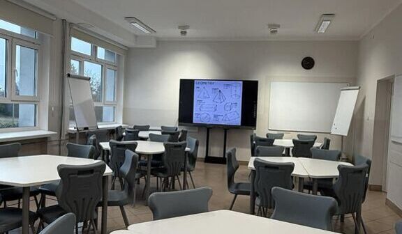 Pusta sala lekcyjna, z sześciokątnymi stolikami i szarymi krzesłami, oświetlona jarzeniówkami. Na środku widoczny ekran z rysunkami figur geometrycznych. Po bokach stoją tablice.