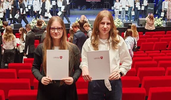 Na zdjęciu widać dwie osoby stojące w teatrze z czerwonymi fotelami, trzymające certyfikaty. W tle scena, na której stoi grupa ludzi w białych koszulach. Na ekranie napis: "Fundusze Europejskie dla Lubelskiego 2021-2027".