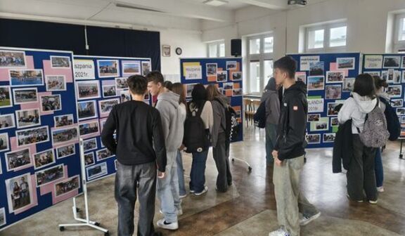 Grupa osób przegląda wystawę zdjęć na stojakach w pomieszczeniu. Tablice oznaczone są nazwami krajów i latami, co sugeruje wystawę podróżniczą lub edukacyjną. W tle jasne okna i odsłonięte sufity.