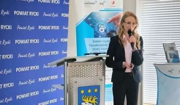 Kobieta w garniturze przemawia przy mównicy z herbem Powiatu Ryckiego. Za nią widoczne są banery z napisami i symbolami. Panuje formalna atmosfera konferencyjna.