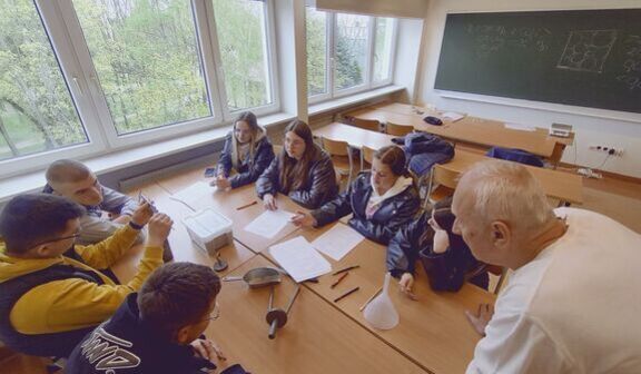 Grupa uczniów siedzi przy stole w klasie, zaangażowana w eksperyment lub projekt. Przed nimi leżą notatki i przybory naukowe. Starszy mężczyzna, prawdopodobnie nauczyciel, patrzy na uczniów.