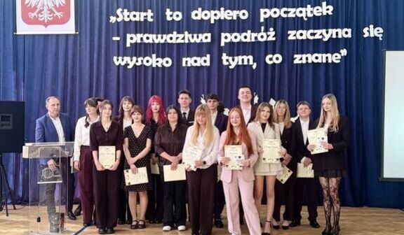 Grupa młodych osób stoi na scenie w auli. W tle niebieska zasłona z polskim godłem i cytatem: "Start to dopiero początek – prawdziwa podróż zaczyna się wysoko nad tym, co znane". Osoby trzymają dyplomy.