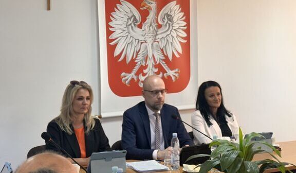 Na zdjęciu troje ludzi siedzących przy stole konferencyjnym, przed nimi mikrofony. W tle widać godło Polski. Obok stoi duża roślina w doniczce.
