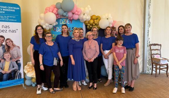 Grupa osób w różnym wieku, głównie kobiety w niebieskich koszulkach, stoi przed dekoracją z balonów. Dekoracja zawiera napis: "Dzień rodziny". Obok stoi stojak z materiałami informacyjnymi.