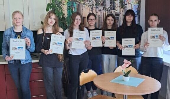 Grupa siedmiu osób, stojących w pomieszczeniu z zielonymi i kwiatowymi dekoracjami, trzyma dyplomy. Na pierwszym planie okrągły stolik z małą rośliną w doniczce.