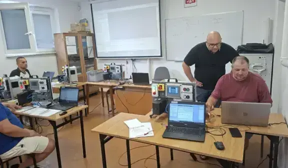 Prowadzący w ciemnej koszulce pochyla się nad stanowiskiem uczestnika w bordowej koszulce i wskazuje coś na ekranie laptopa. Na stole dwa laptopy i moduł sterowniczy z panelem. W tle kolejni uczestnicy i ekran projekcyjny.