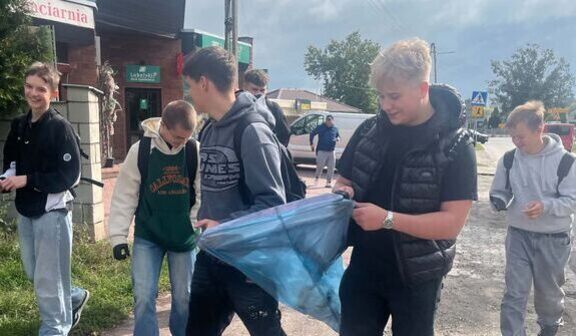 Grupa nastolatków na chodniku z czerwonej kostki; dwóch chłopców trzyma rozłożony niebieski worek na śmieci, pozostali stoją obok. W tle pawilon handlowy ze spiczastym dachem, drzewa i zachmurzone niebo.