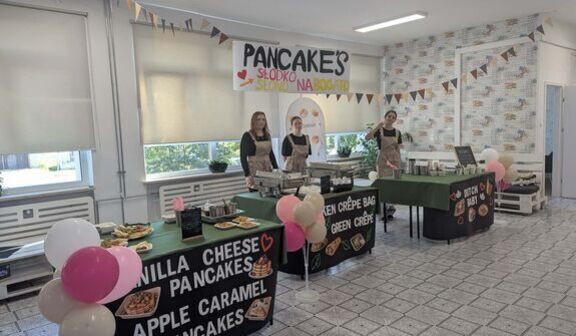 Sala z jasną posadzką i stoiskami nakrytymi zielonymi obrusami, z czarnymi banerami z napisami Vanilla Cheese Pancakes, Apple Caramel Pancakes, Chicken Crêpe Bag i Green Crêpe. Przy pierwszym stoisku pęk różowych i białych balonów, na ścianie kolorowy napis Pancakes i girlandy z chorągiewek, w głębi trzy osoby w fartuchach.
