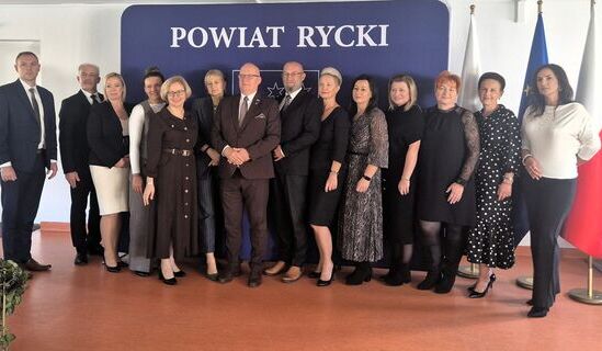Grupa około dwudziestu nauczycieli, pracowników oświaty i przedstawicieli władz powiatu stoi w sali przy granatowej ściance z napisem Powiat Rycki, obok flagi Polski i Unii Europejskiej.