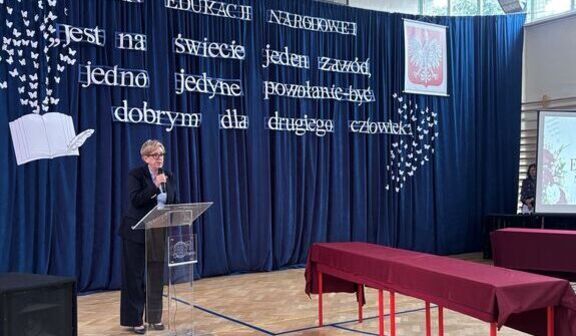Kobieta przemawia przy przezroczystej mównicy w szkolnej sali gimnastycznej. Za nią granatowa kotara z napisem: Dzień Edukacji Narodowej. Jest na świecie jeden zawód, jedno jedyne powołanie – być dobrym dla drugiego człowieka. Obok godło Polski.