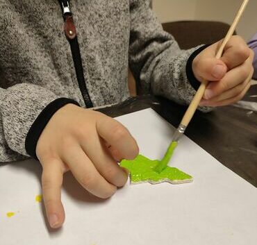 Dziecko maluje zieloną farbą na białej kartce, trzymając pędzel w prawej dłoni. Widać tylko ręce dziecka.