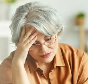 Kobieta z siwymi włosami w okularach przyciska dłoń do czoła w geście stresu lub zmęczenia, ubrana w pomarańczową bluzkę, na rozmytym tle.