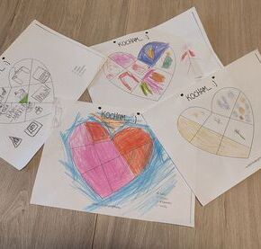 Rysunki dzieci przedstawiające kolorowe serca na kartkach rozłożone na drewnianej podłodze.