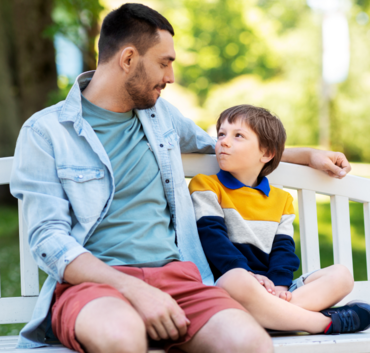 Mężczyzna i chłopiec siedzą na ławce w parku, zwróceni do siebie, obydwoje uśmiechnięci i wydają się być w rozmowie.