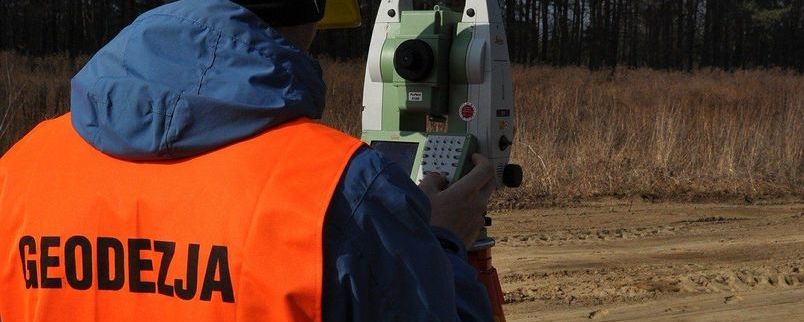 mężczyzna w pomarańczowej kamizelce robiący pomiary geodezyjne w terenie