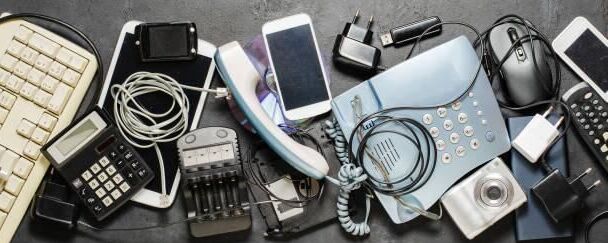 Zbiór różnych urządzeń elektronicznych na ciemnym tle: klawiatura, kalkulator, telefony, ładowarki, aparat, mysz komputerowa. Przedmioty są ułożone chaotycznie, ukazując różnorodność technologii.