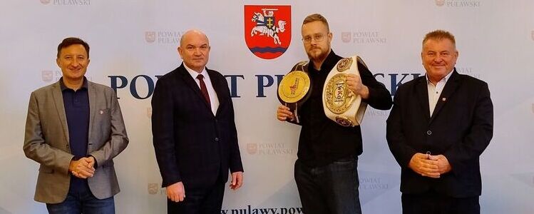 Spotkanie z mistrzem świata Jackiem Błasińskim w Starostwie Powiatowym w Puławach. Ludzie na tle ścianki z herbem Powiatu Puławskiego.