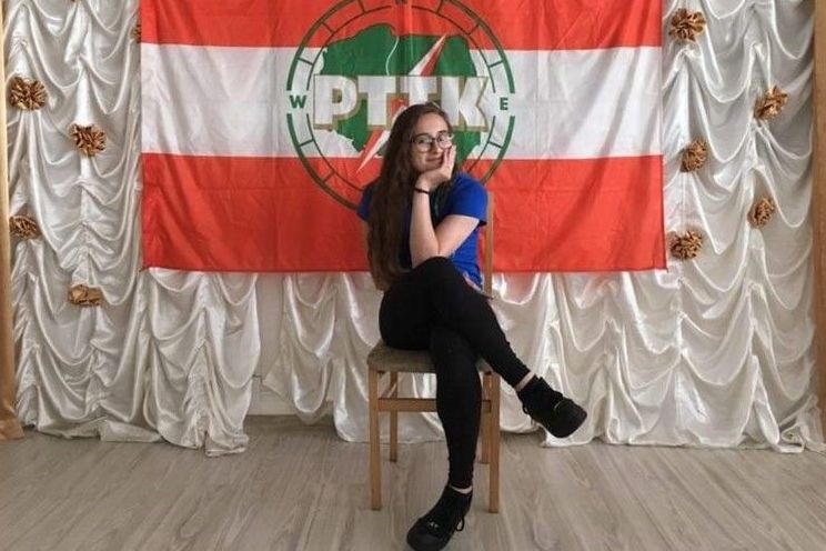 Na zdjęciu  dziewczyna siedząca na krześle na tle flagi PTTK