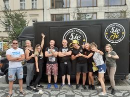 Zdjęcie przedstawia roześmianą załogę na tle ich food trucka. Samochód ma kolor czarny, napisy na nim są białe. W tle widać dawny dom towarowy. 