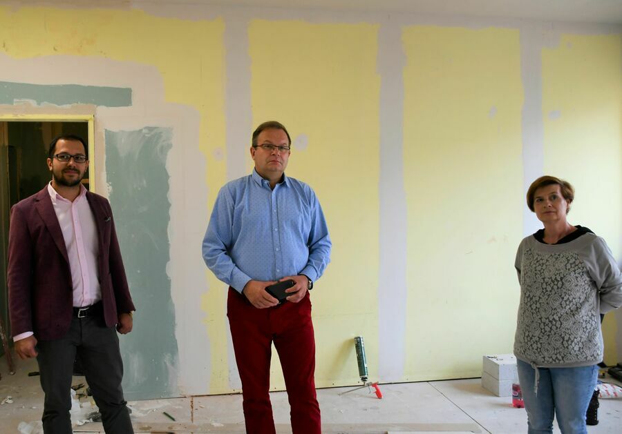 Na zdjęciu widoczne trzy osoby: dwóch mężczyzn i jedna kobieta, stojące w remontowanej sali o żółtych ścianach. 