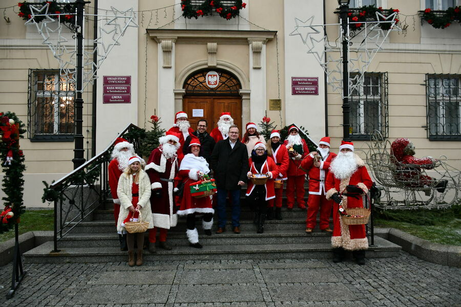 Na zdjęciu widać Burmistrza Miasta oraz Zastępcę w otoczeniu osob w strojach Mikołajek i Śnieżynek stojących na schodach prowadzących do Ratusza.