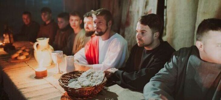 Grupa mężczyzn siedzi przy stole, na którym znajdują się chleb i dzban, przywodzące na myśl scenę z Ostatniej Wieczerzy. Centralna postać ma na sobie białą szatę i czerwoną przepaskę.