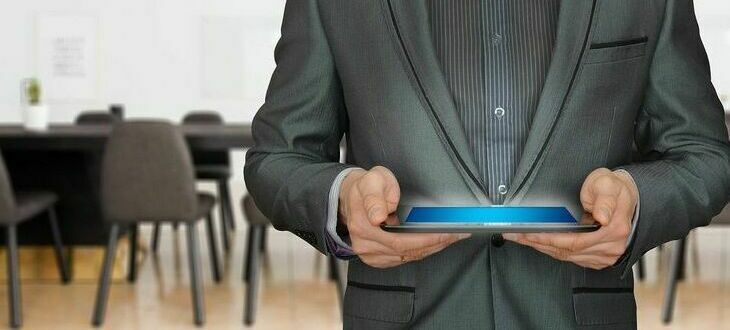 Osoba w garniturze trzyma poziomo tablet, wyświetlacz świeci niebieskim kolorem. Tło to rozmyte biuro.