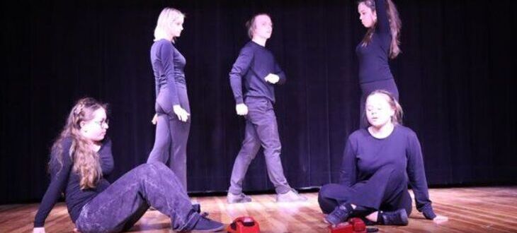 Na zdjęciu jest pięcioro ludzi na scenie, którzy wydają się przygotowywać do występu lub ćwiczyć choreografię. Noszą casualowe, ciemne stroje i mają skupione wyrazy twarzy.