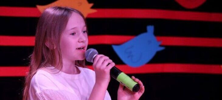 Młoda dziewczyna śpiewająca do mikrofonu z skupionym wyrazem twarzy. Tło sceny zdobią abstrakcyjne formy w czerwono-czarnych barwach.