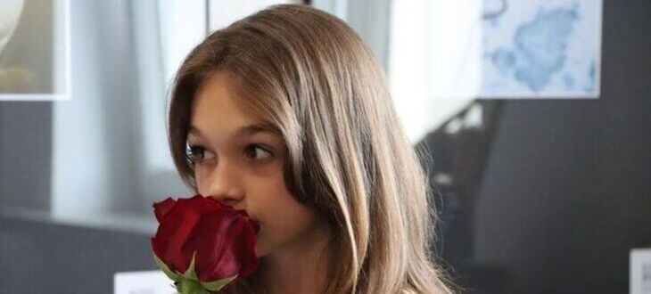Młoda dziewczyna z włosami do ramion wącha czerwoną różę, stojąc w pomieszczeniu z sztuką na ścianach.