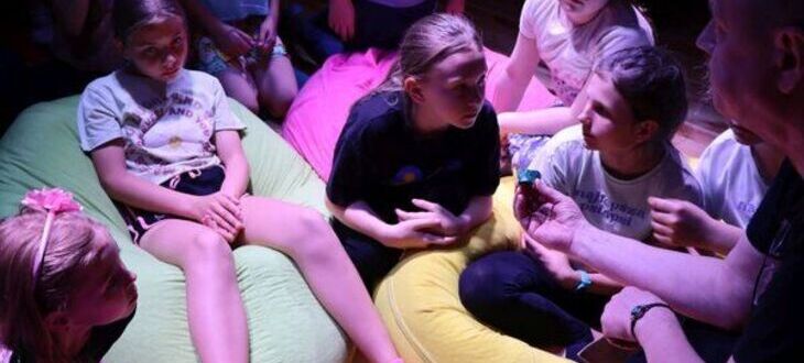 Grupa dzieci siedzi na kolorowych poduszkach w półokręgu, wydają się być zaangażowane w rozmowę lub aktywność grupową.