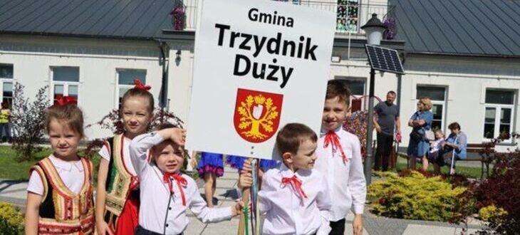 Grupa dzieci w tradycyjnych polskich strojach ludowych trzyma tablicę z napisem "Gmina Trzydnik Duży" i herbem, stoją przed budynkiem.