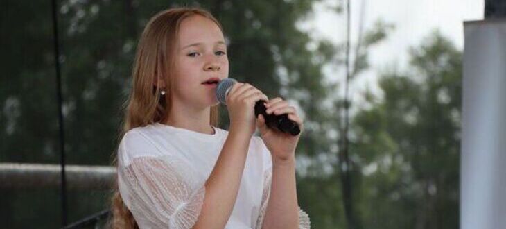 Młoda dziewczyna śpiewa do mikrofonu na scenie na zewnątrz, ubrana w biało-różową sukienkę, w tle widoczne drzewa i banery.