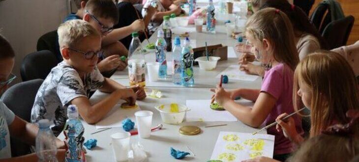 Grupa dzieci siedzi przy długich stołach, malując i biorąc udział w zajęciach plastycznych. Na stole widać farby, pędzle i butelki z wodą.