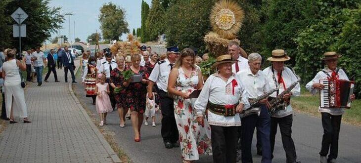 Procesja ludzi ubranych w tradycyjne stroje, z muzykami z akordeonami na czele, idąca ulicą wsi.