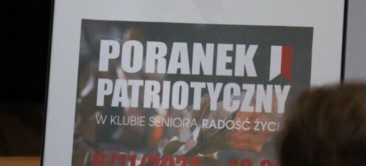 Na zdjęciu widoczna jest drewniana figurka - młotek umieszczony na tablicy z napisem "Poranek Patriotyczny w Klubie Seniora Radość Życia". Pod młotkiem znajduje się czerwona róża.